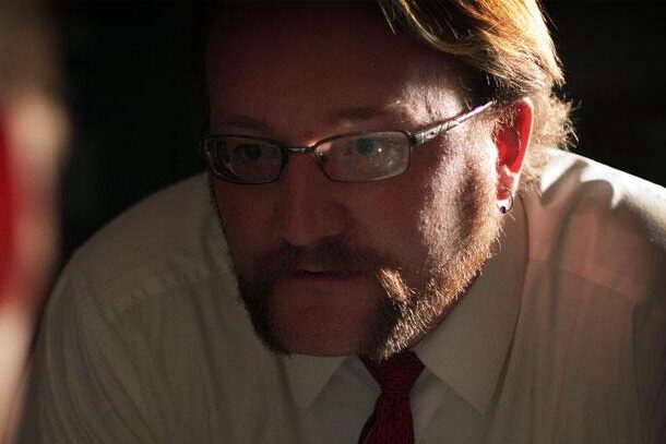 a still from the movie Downcast, featuring lead actor Ian Herrmann in dark noir-style lighting, his grim expression as the character Harry interrogates his former subordinate